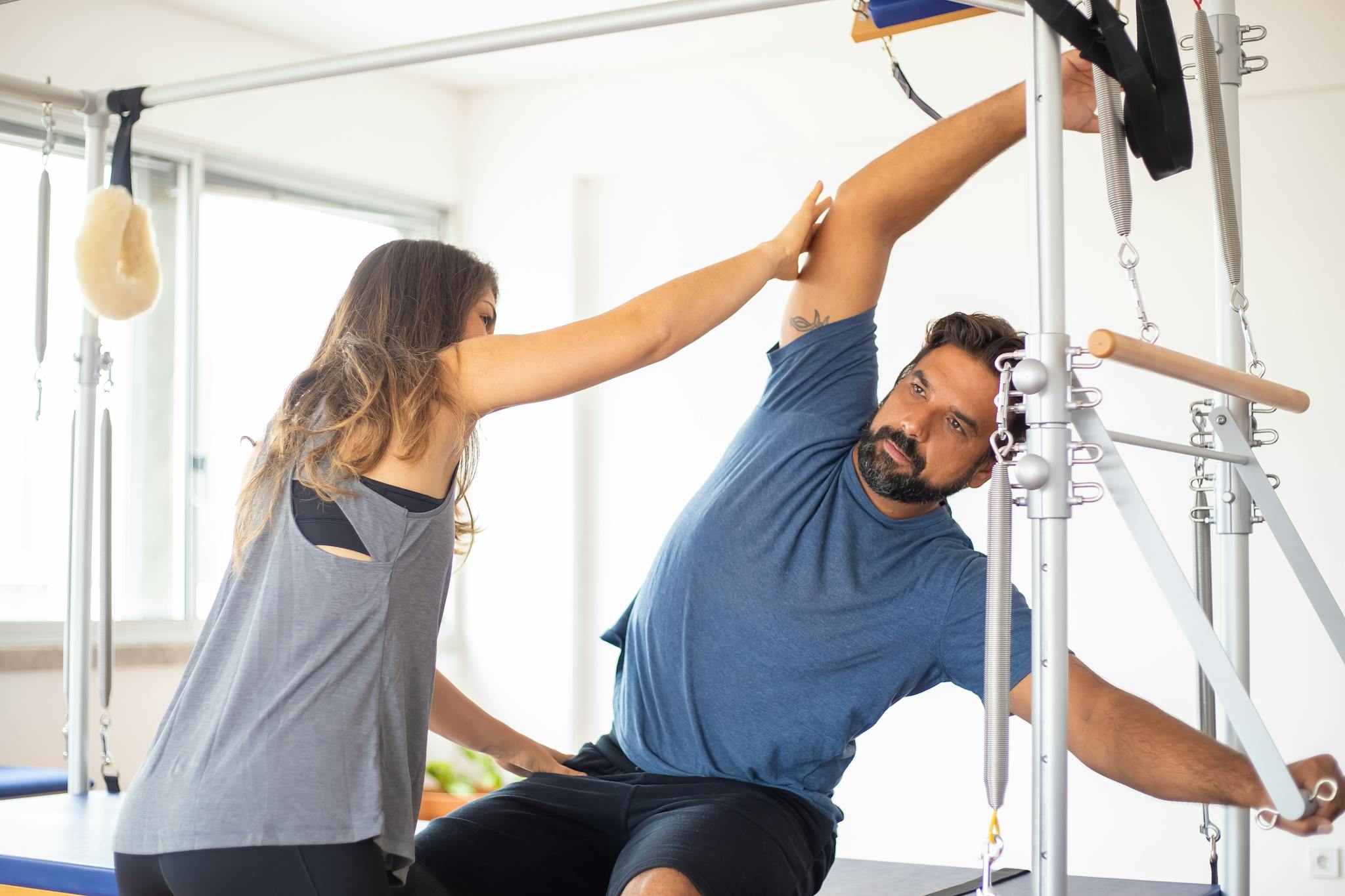 Trainer guides client on reformer machine during pilates session in a bright gym setting.